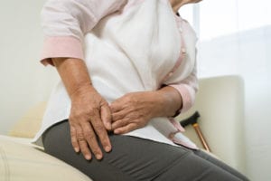 elderly woman sitting on the side of the bed leaning over and clutching her hip in pain.