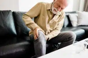 A distraught senior man feeling unwell, suffering from pain in leg while sitting on sofa in the living room at home, Photo of elderly man sitting on a sofa in the living room at home and touching his knee by the pain during the day. Senior man massaging his painful knee