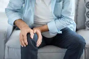 Older man who can benefit from knee replacement surgery sitting on a couch and gripping his knee in discomfort.